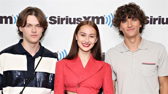 The image shows three young people standing side by side in front of a white backdrop with the 